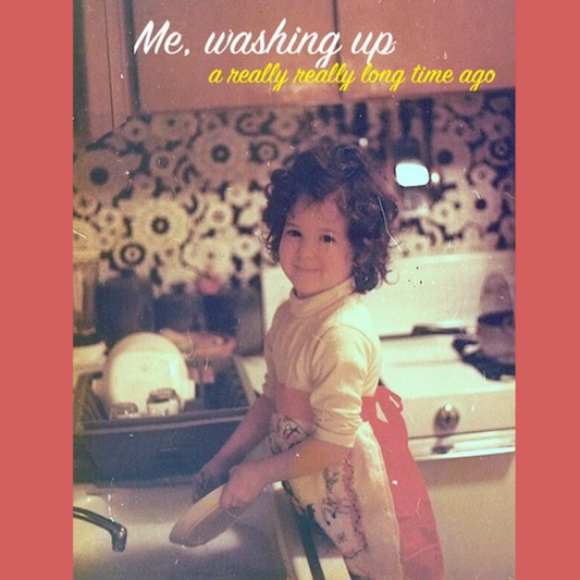 A little girl with curly dark hair washes dishes at the sink in a 1970s kitchen