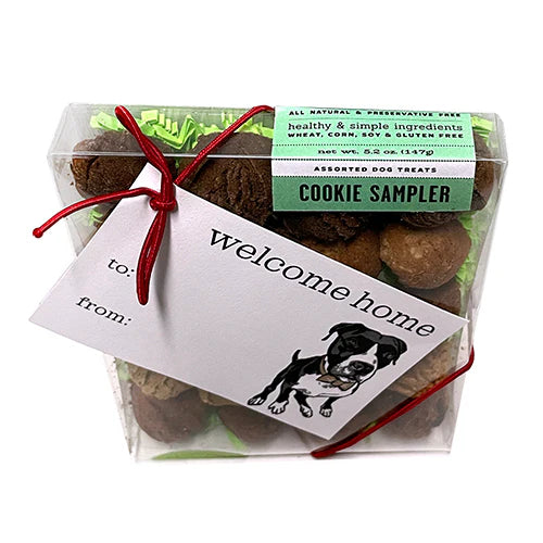 Top view of twenty round basked dog cookies inside a clear plastic box with green paper grass and a welcome home gift tag portraying a black and white pit bull-type dog 