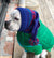 A white pit bull-type dog sitting down wearing a blue knitted hat with pom pom and a green parka