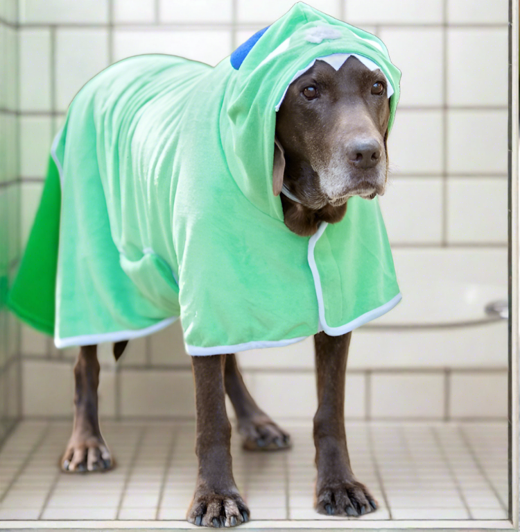 a large brown dog wearing a green Terry Bath Robe with a Monster face and horns on the hood, standing in a white tiled shower